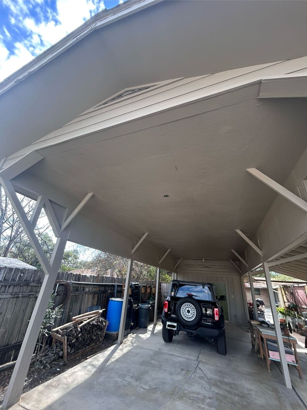 Carport with gray ceiling, parked SUV, and backyard storage area. Bright cloudy sky visible.