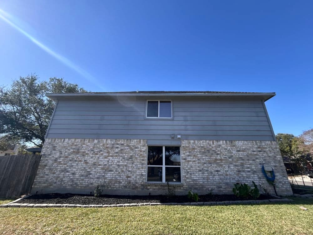 Two-story residential home with gray siding and brick foundation under a clear blue sky.